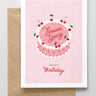 A greeting card with a pink background featuring a graphic of a cake with cherries on top and the text 'Forever Young' in a circular pattern. The card also includes a 'Happy Birthday' message and comes with a brown envelope.