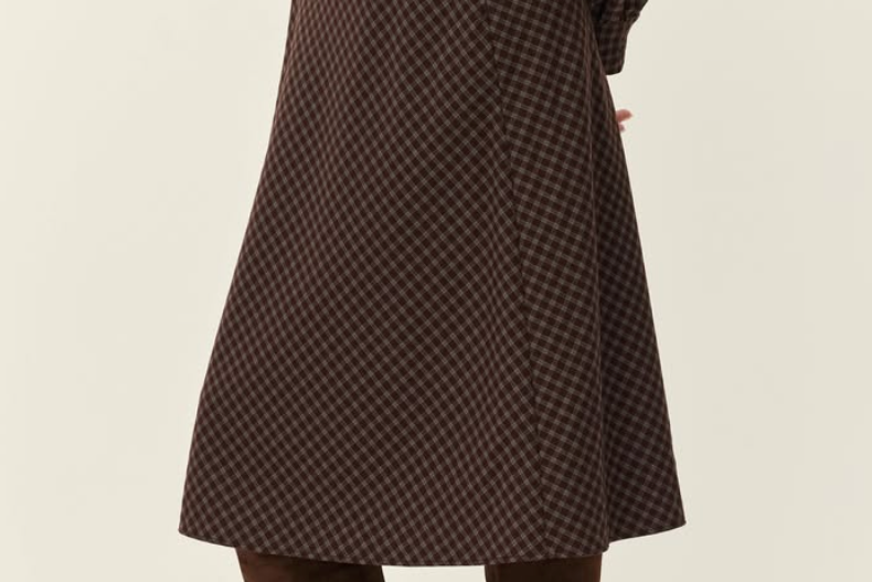 A person shown from the back wearing a brown and black checkered midi dress with long sleeves and a matching belt, paired with brown suede over-the-knee boots, against a plain light beige studio background.