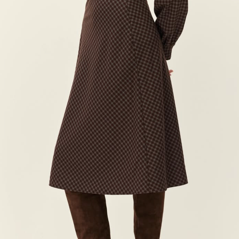 A person shown from the back wearing a brown and black checkered midi dress with long sleeves and a matching belt, paired with brown suede over-the-knee boots, against a plain light beige studio background.