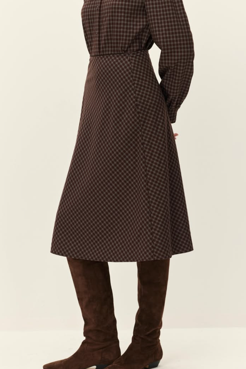 A person shown from the back wearing a brown and black checkered midi dress with long sleeves and a matching belt, paired with brown suede over-the-knee boots, against a plain light beige studio background.