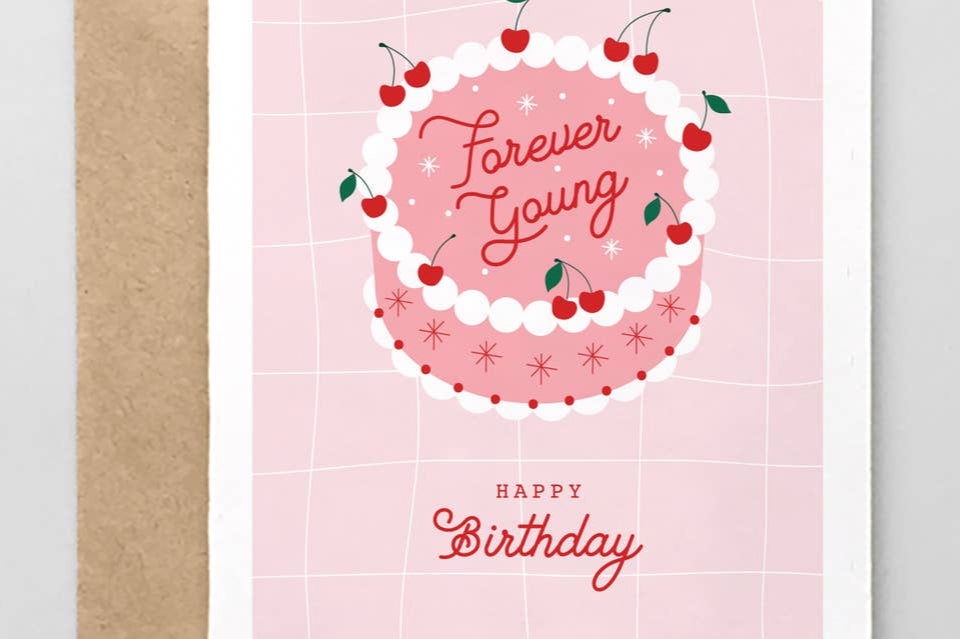A greeting card with a pink background featuring a graphic of a cake with cherries on top and the text 'Forever Young' in a circular pattern. The card also includes a 'Happy Birthday' message and comes with a brown envelope.