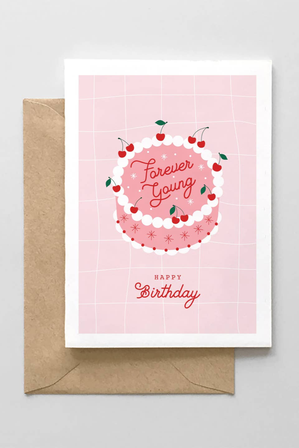 A greeting card with a pink background featuring a graphic of a cake with cherries on top and the text 'Forever Young' in a circular pattern. The card also includes a 'Happy Birthday' message and comes with a brown envelope.